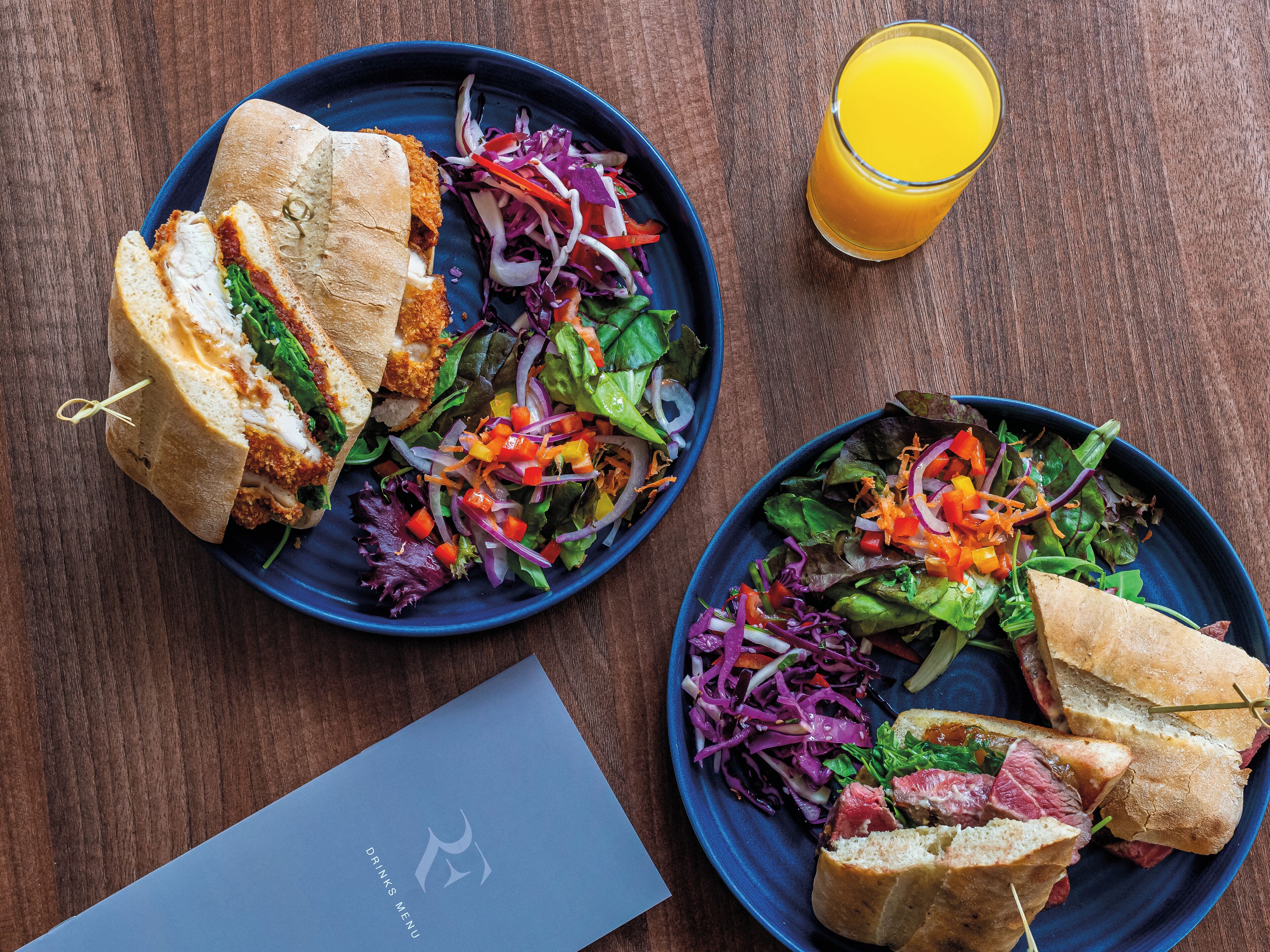 A meal consisting of sandwiches and salads on blue plates, a glass of orange juice, and a menu on a wooden table.