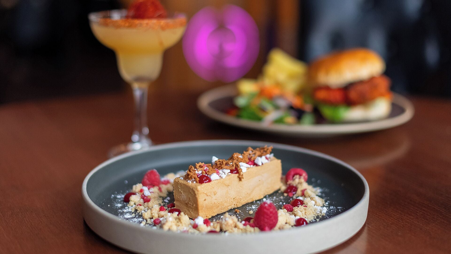 A dessert plate with a layered cake, garnished with berries and crumbs, and a cocktail glass in the background.