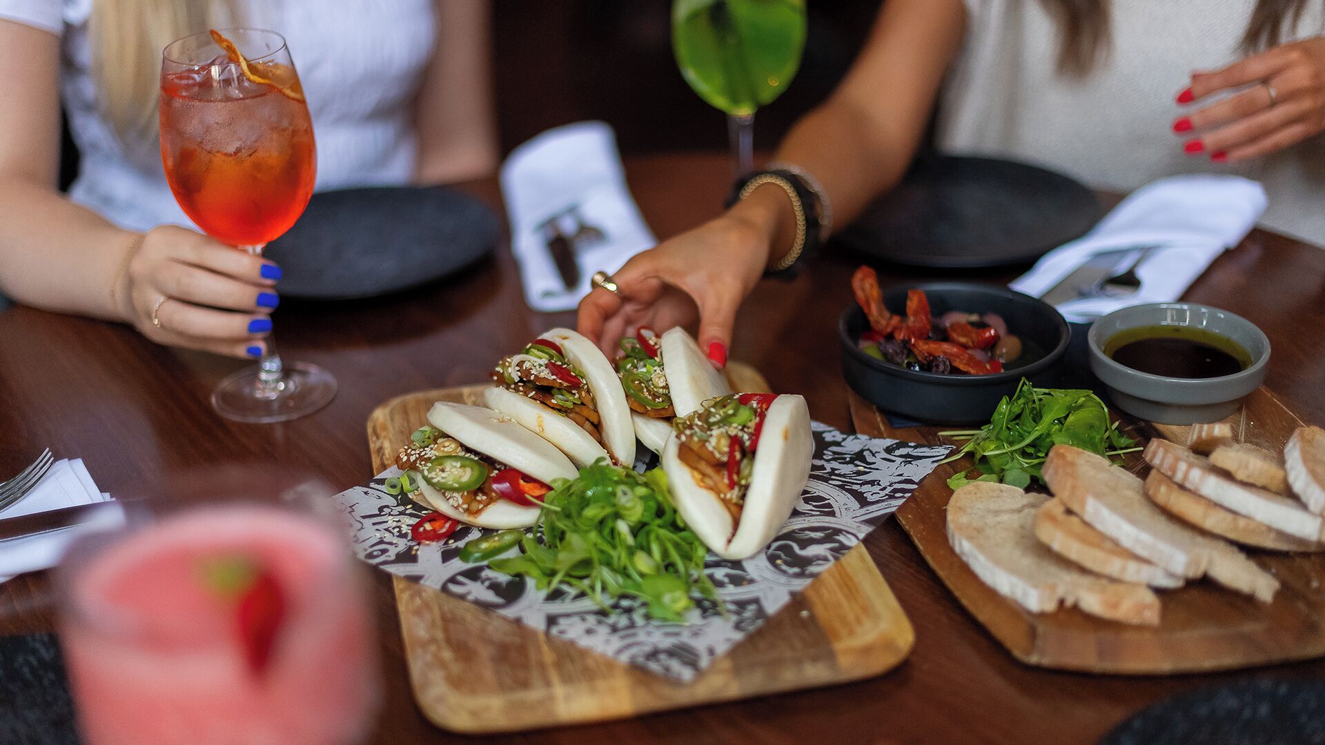 Una mesa de comedor con comida y bebidas, incluyendo un plato con carne y vegetales en rodajas, un bol de aceitunas y un vaso de bebida roja.