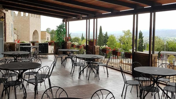 An outdoor patio area with round tables and chairs, a wooden ceiling, and a view of greenery and a distant landscape. - Hotel il Castello (Assisi)