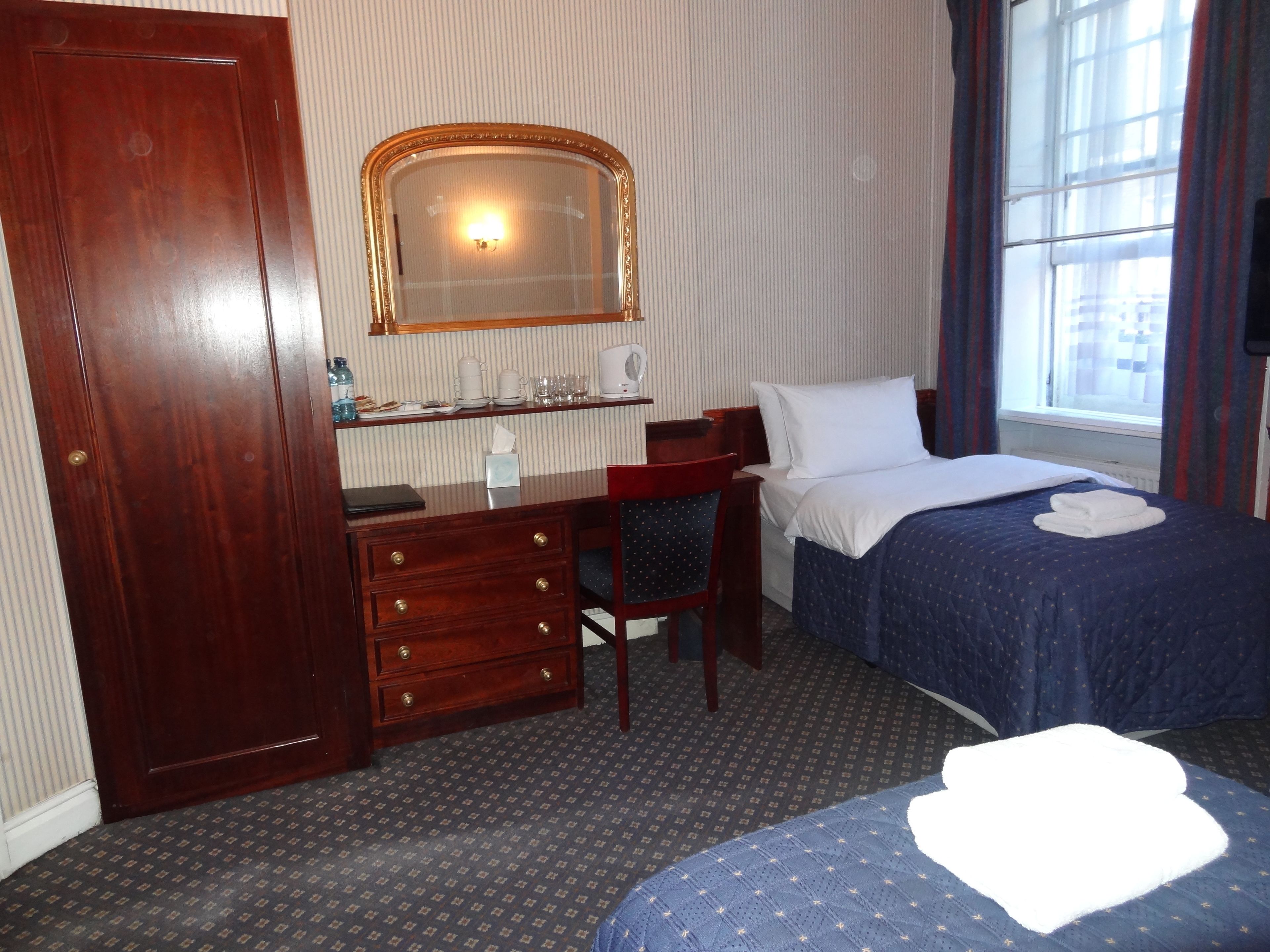 Habitació d'hotel amb escriptori de fusta, una cadira, un llit amb col·cha blava, una taula de nit, un mirall i una finestra amb cortines.