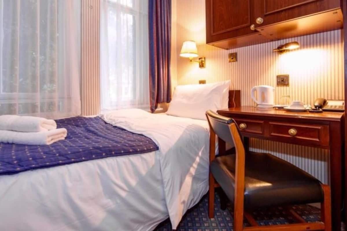 A hotel room with a bed, a desk, a chair, a lamp, and a window with curtains.