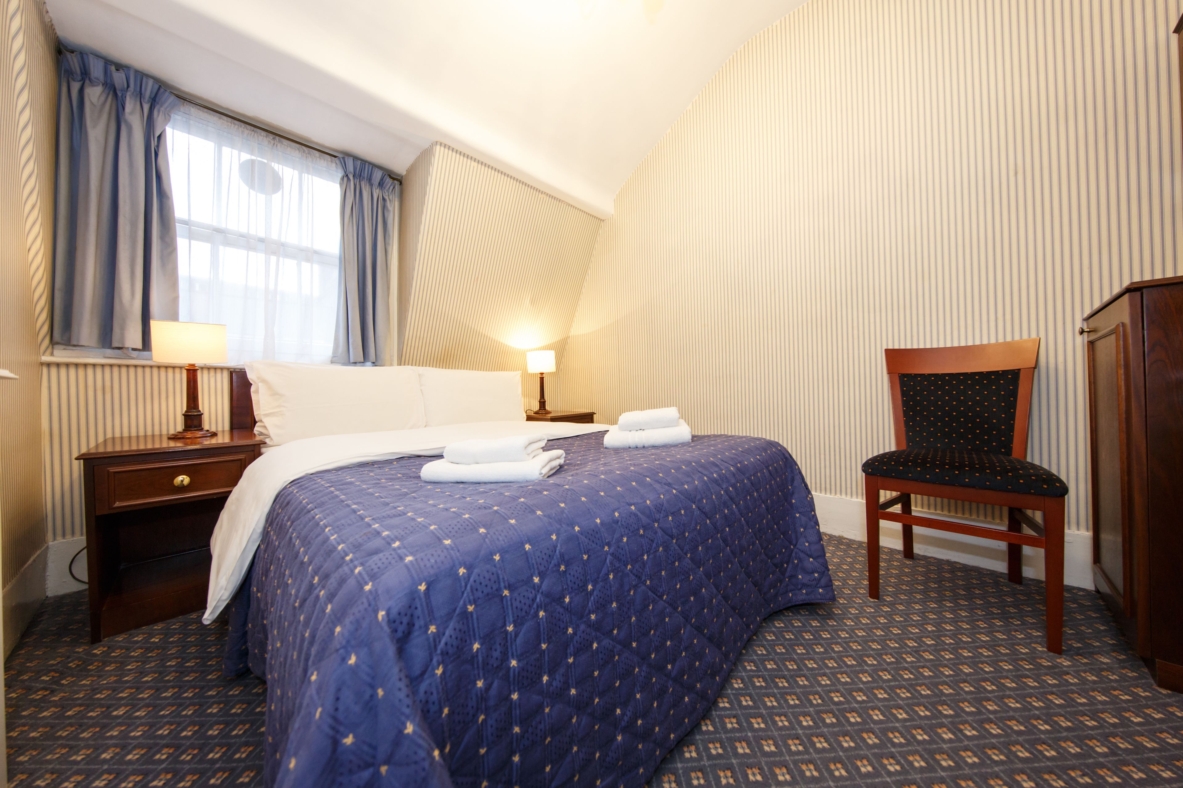 A hotel room with a neatly made bed, a chair, a nightstand with a lamp, and a window with curtains.