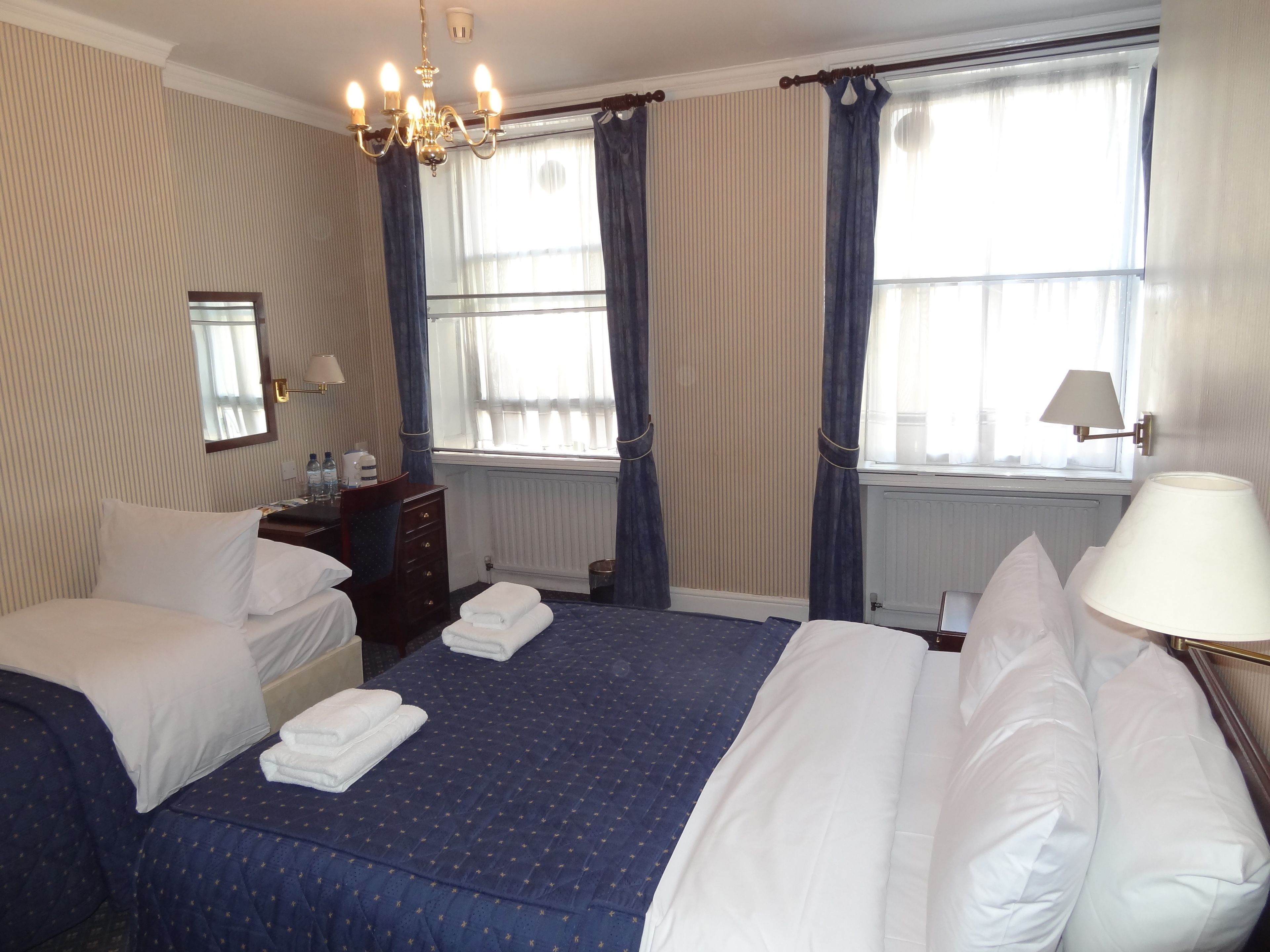 Une chambre d’hôtel avec deux lits, un lustre, une commode, un miroir et une fenêtre avec des rideaux.