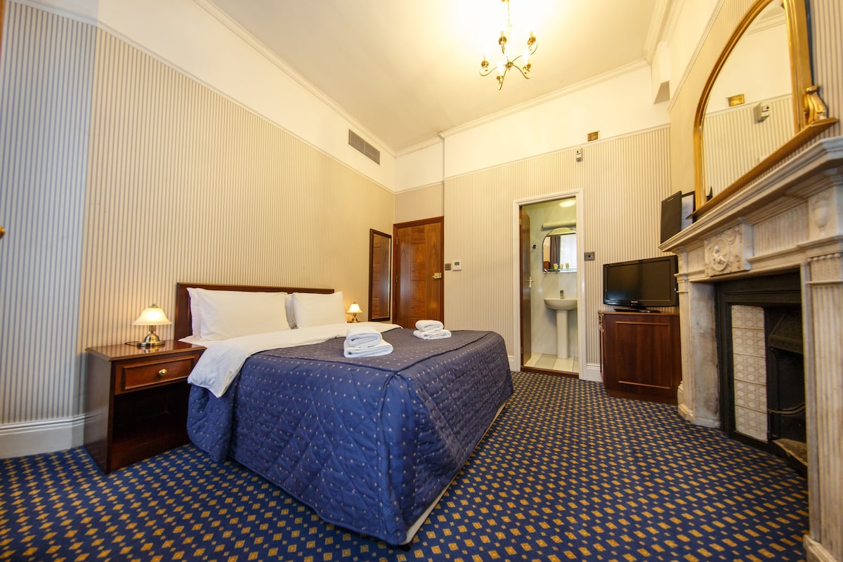 A hotel room with a large bed, a television, a fireplace, and a mirror.