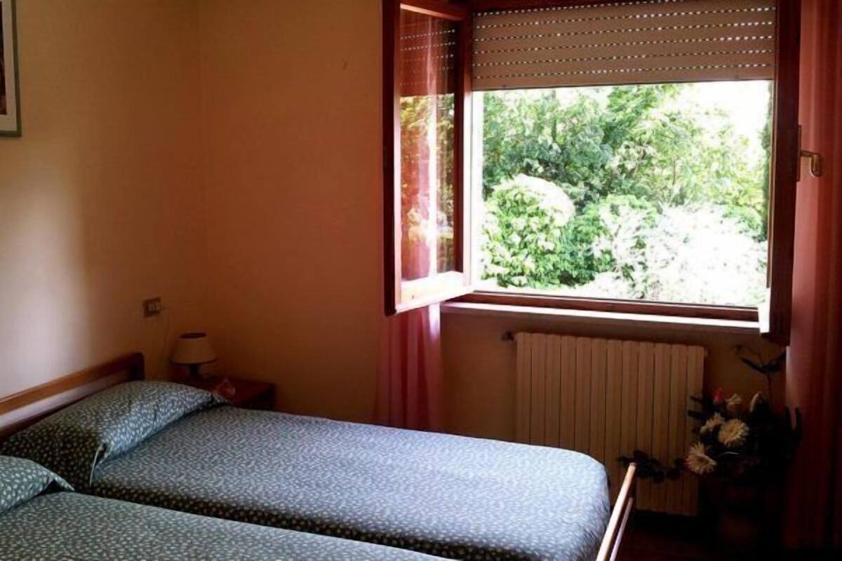 A room with two beds, a window with a view of greenery, a radiator, and a small table with a lamp.