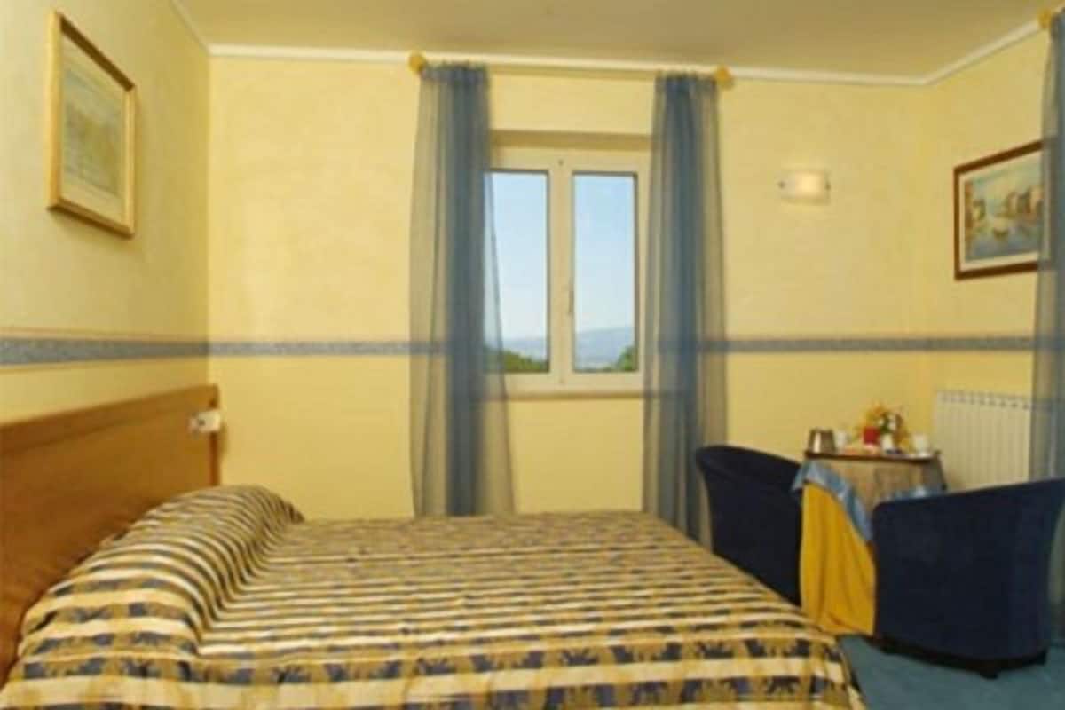 A hotel room with a bed, a small table with a chair, and a window with curtains.