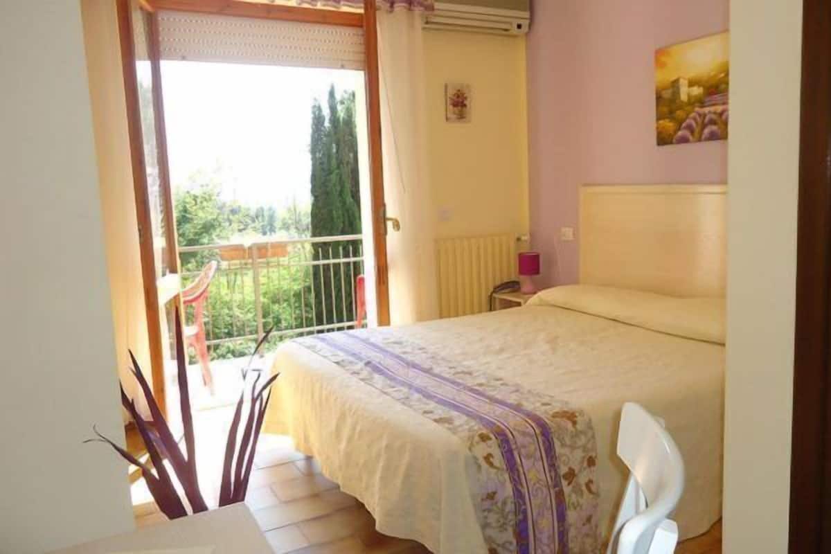 A bedroom with a bed, a chair, a table, a radiator, and a window with a view of trees.