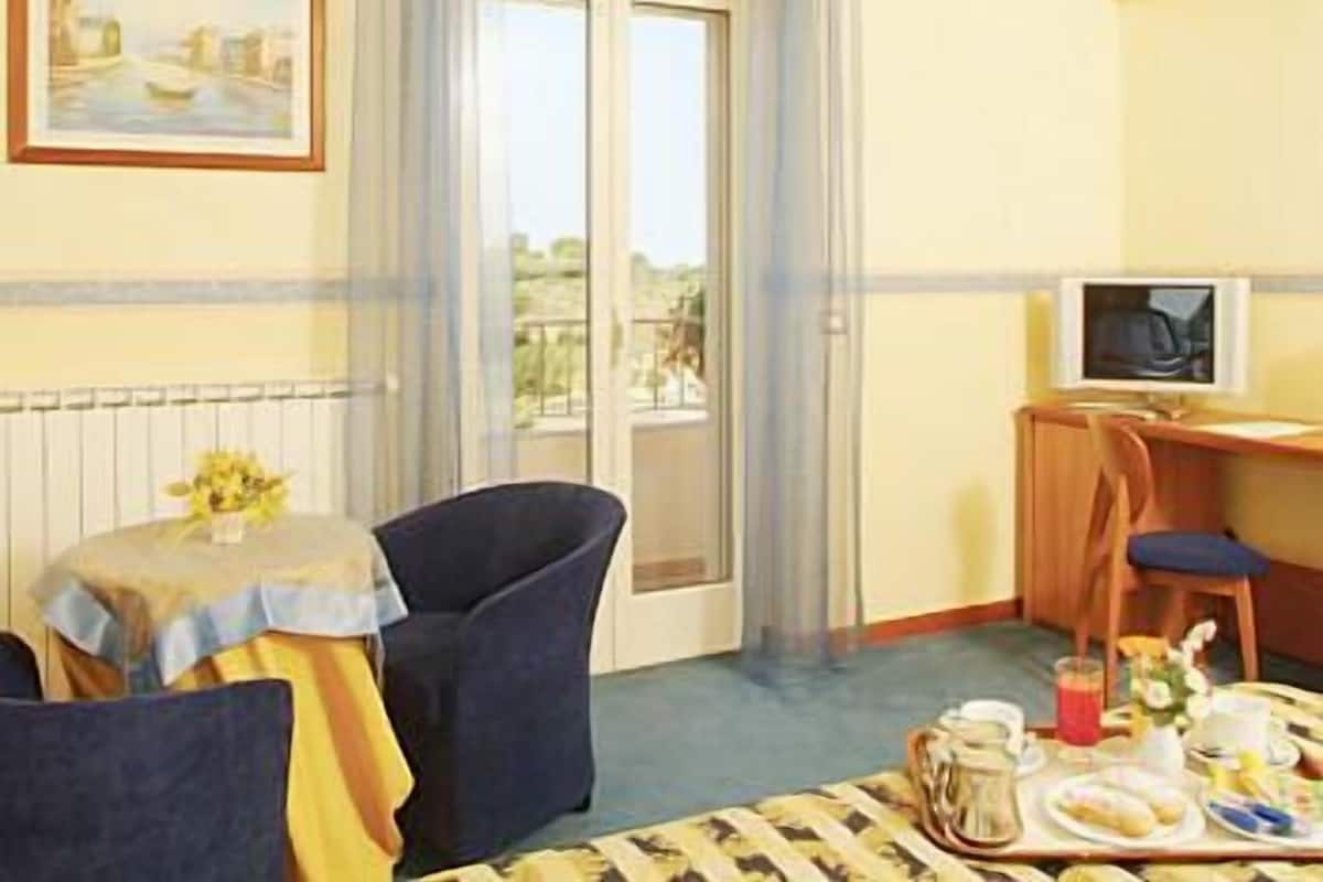 A room with a dining table set for breakfast, a chair, a small table with a vase, and a painting on the wall.