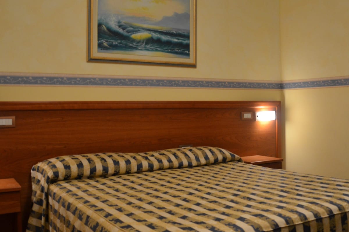 A hotel room with a large bed, a wooden headboard, a painting of a seascape, and a nightstand with a lamp.