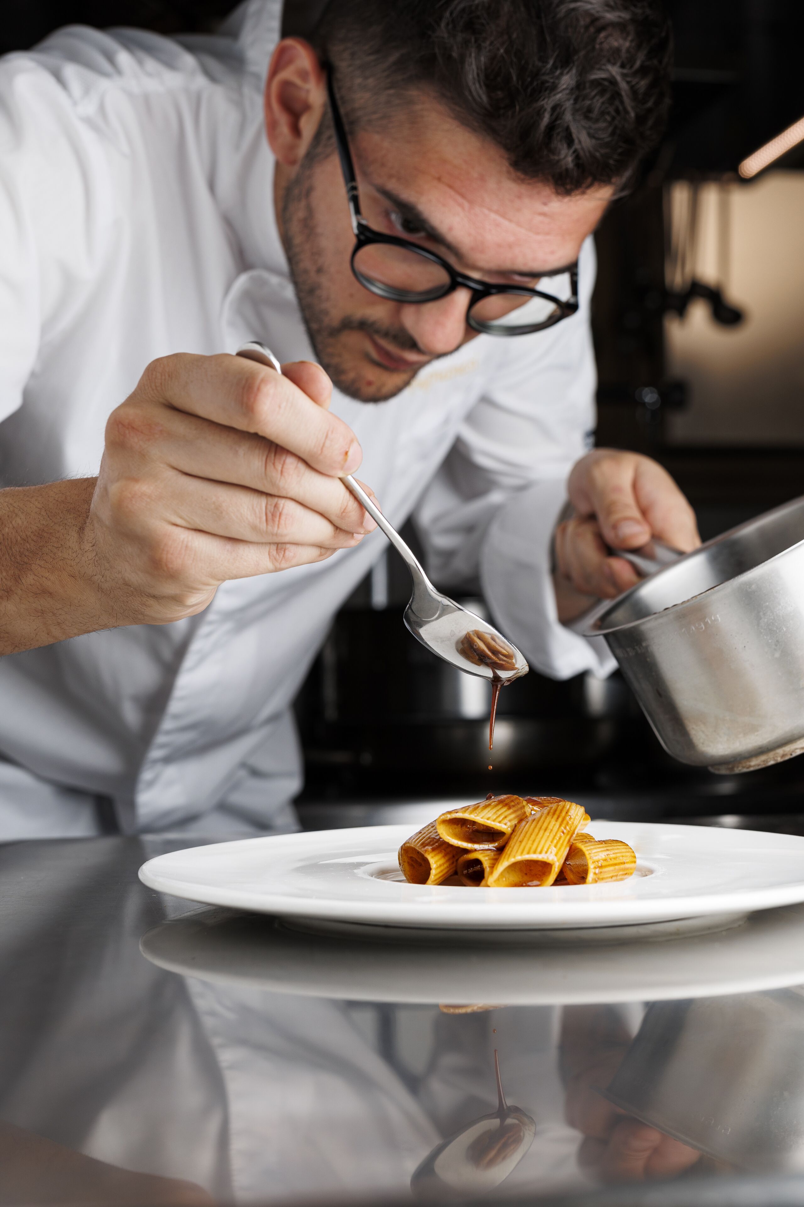A person in a white chef's coat pouring sauce from a pot onto a plate of pasta.