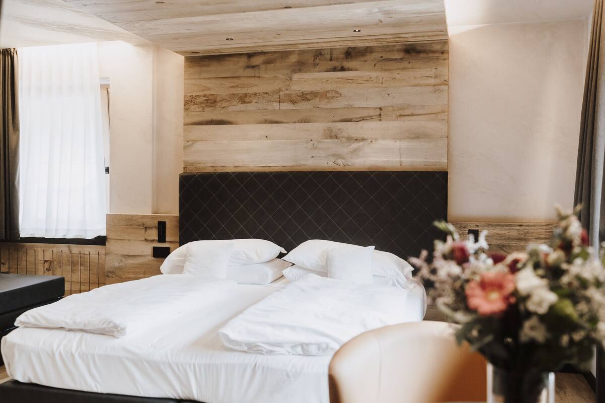 Habitación de hotel con cabecera de madera, cama con ropa blanca, mesita de noche y ventana con cortinas.
