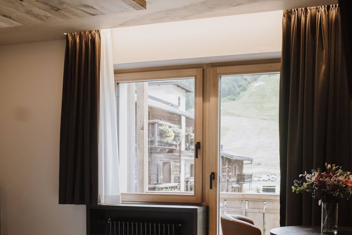 Un dormitorio con cama, una silla, una mesa con un florero y una ventana con cortinas.