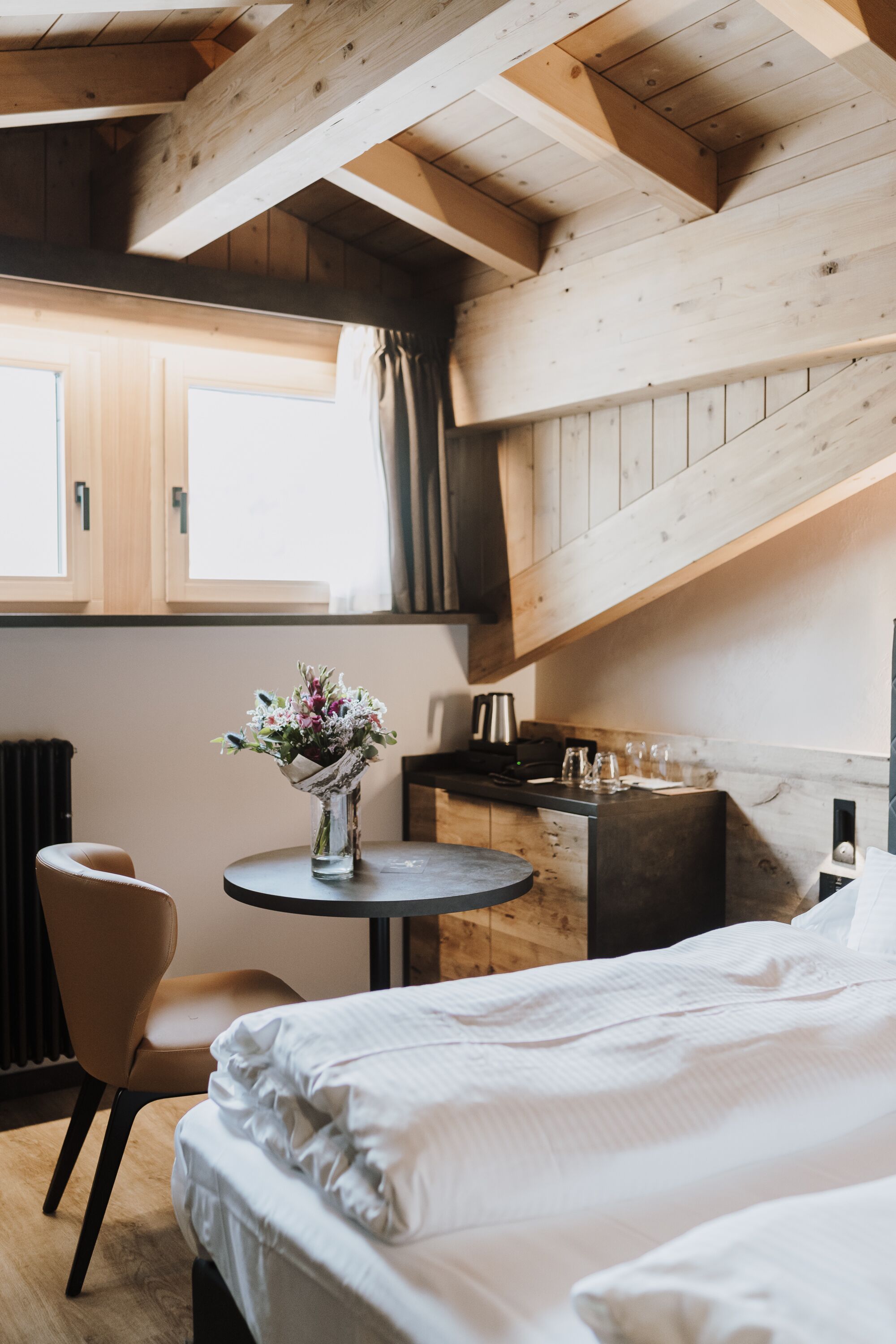 A bedroom with a bed, a small round table with a vase of flowers, a chair, and a kitchenette area.