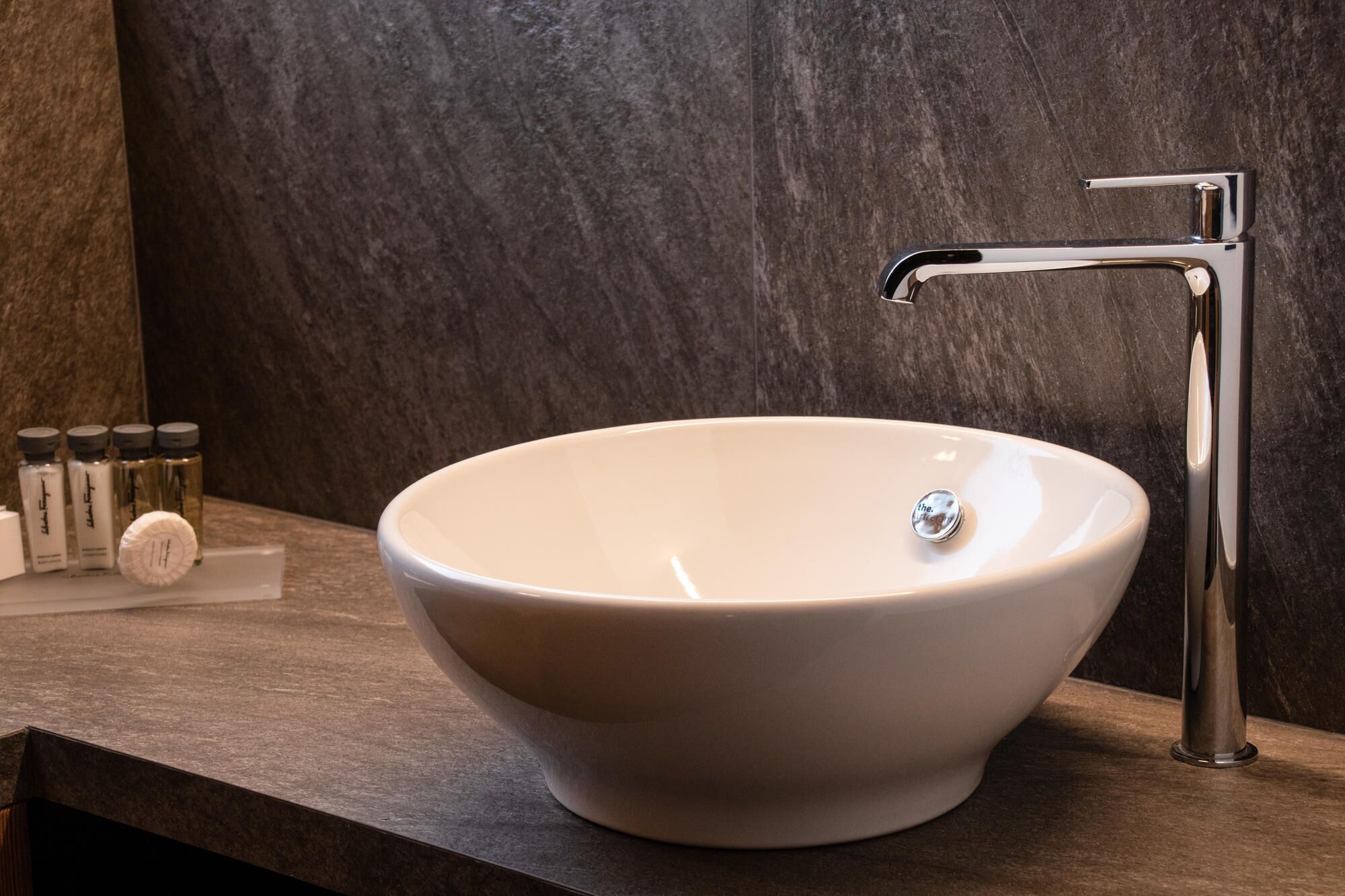 Un baño moderno con un lavamanos blanco tipo recipiente, un grifo cromado y una pared oscura con textura.