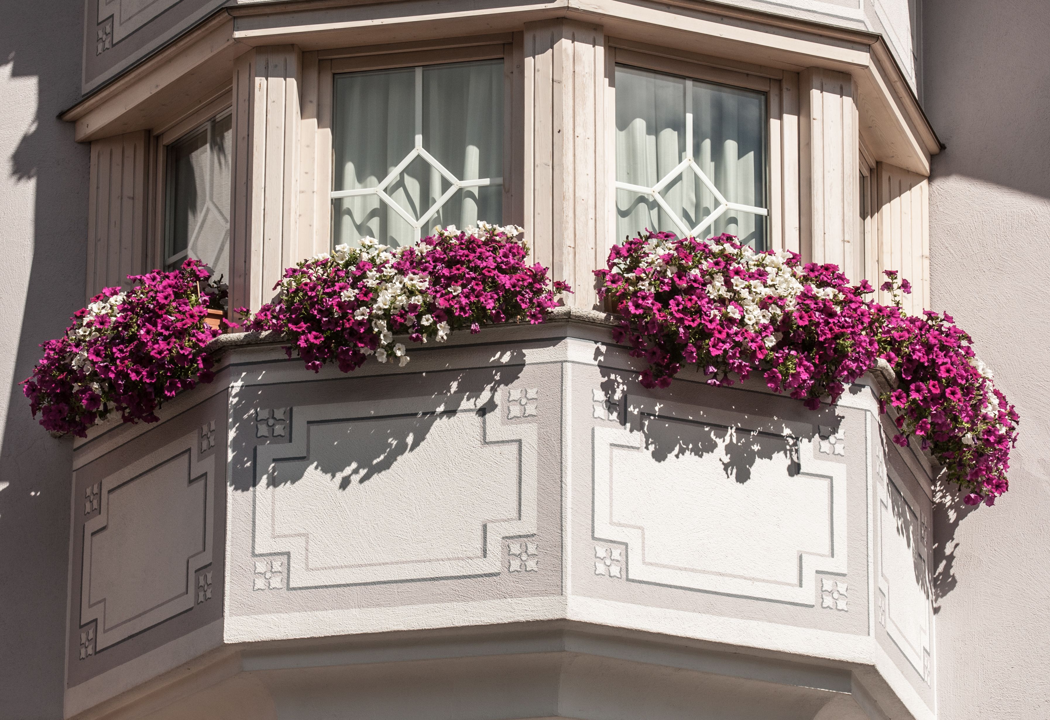 Een raam op de hoek met decoratieve luiken en bloembakken met roze en witte bloemen.