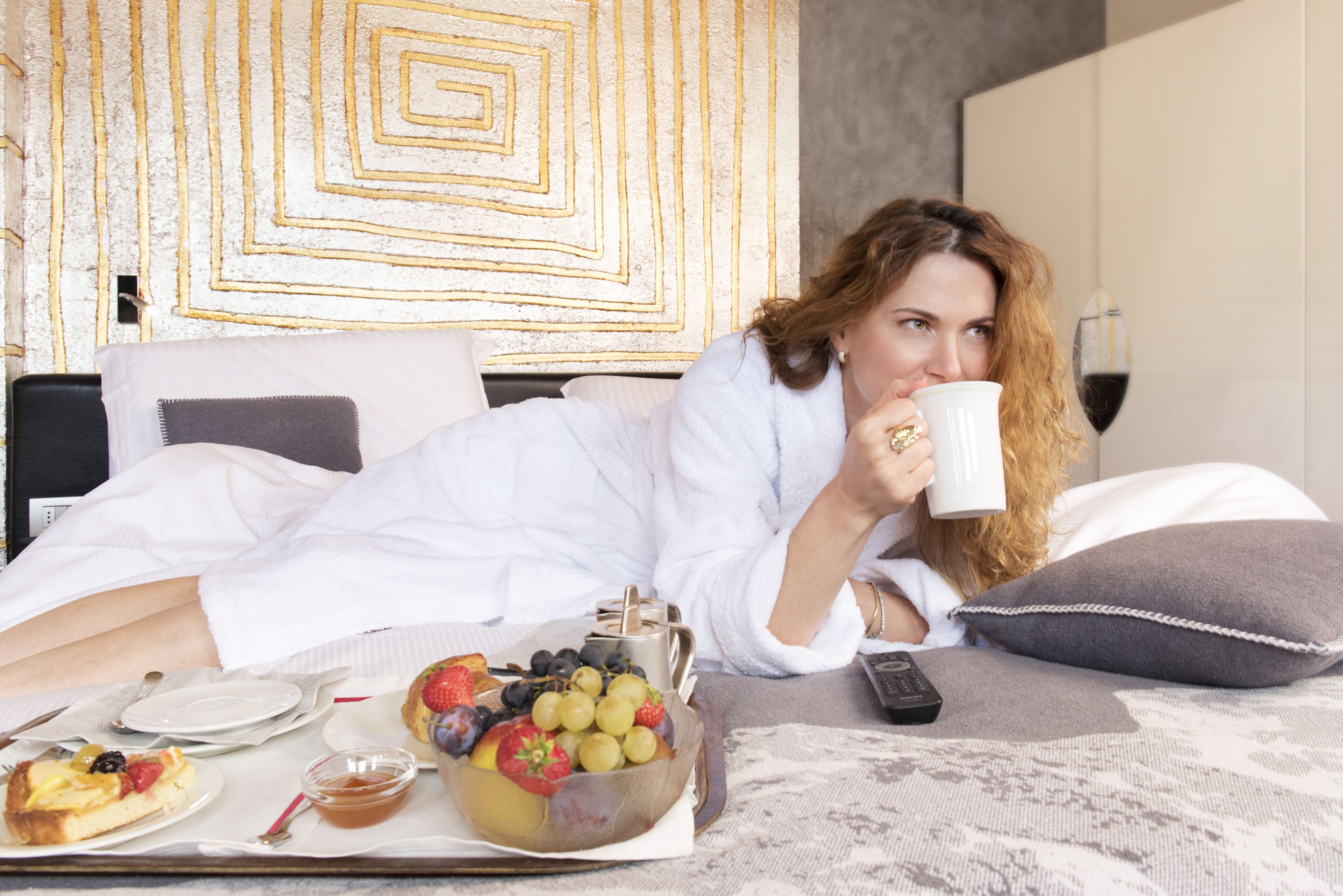 A person lying on a bed with a tray of breakfast items, including toast, fruit, and a drink.