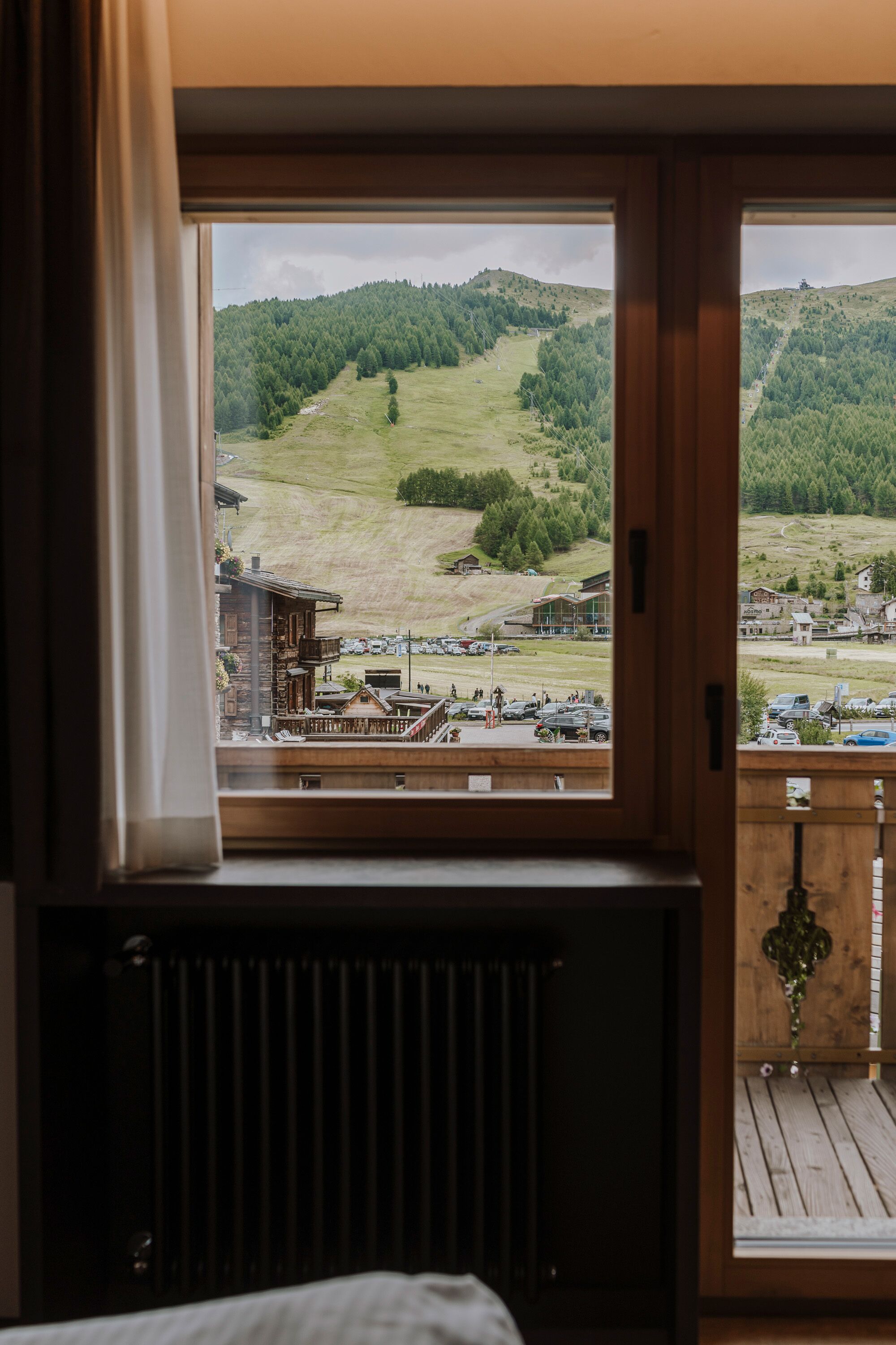 Ein Zimmer mit großem Fenster und Blick auf eine gebirgige Landschaft, mit Holzbalkon und Heizkörper.