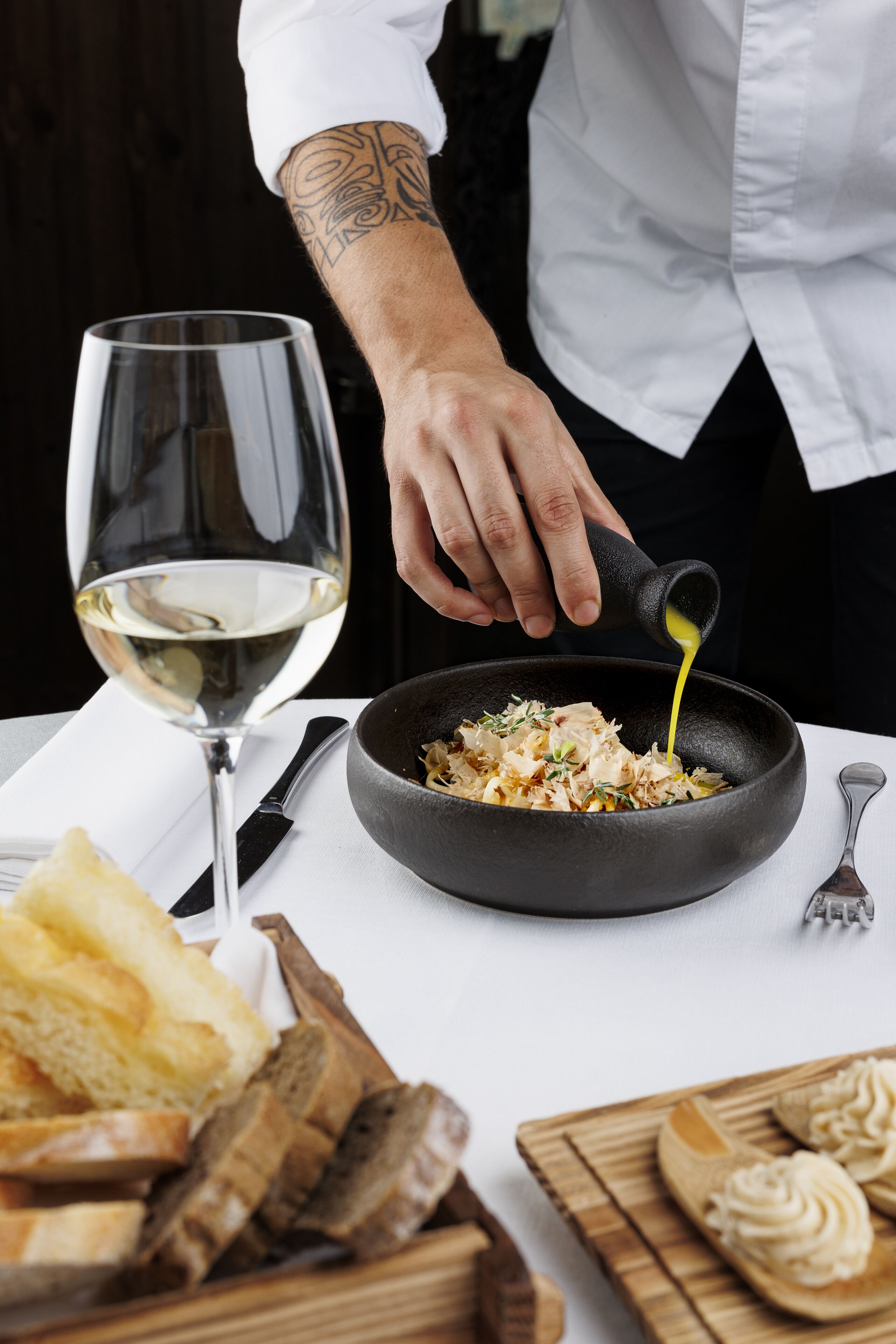 A chef pouring sauce into a pan of food, with a glass of white wine and bread on the table.