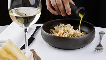 A chef pouring sauce into a pan of food, with a glass of white wine and bread on the table.