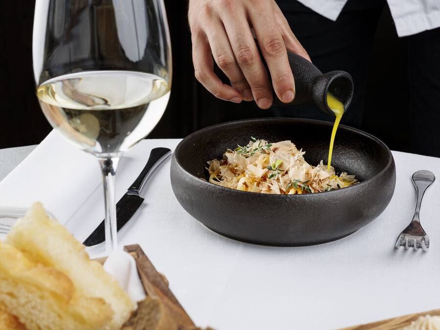 A chef pouring sauce into a pan of food, with a glass of white wine and bread on the table.