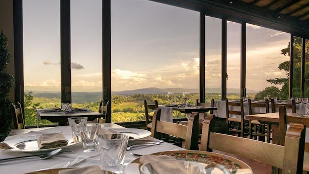 Un área de comedor al aire libre con mesas dispuestas para dos, con vistas a un paisaje pintoresco con montañas y árboles.
