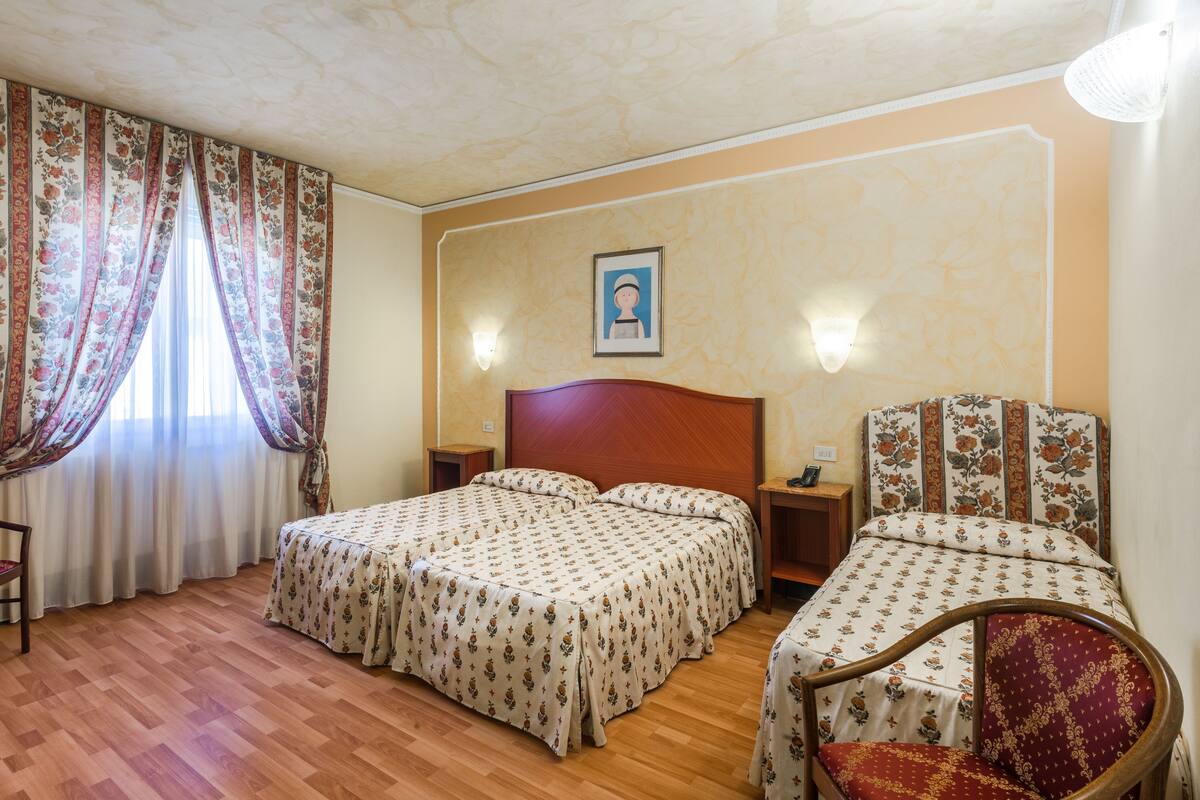 A hotel room with two beds, a wooden floor, floral curtains, a framed picture on the wall, and a nightstand with a phone.