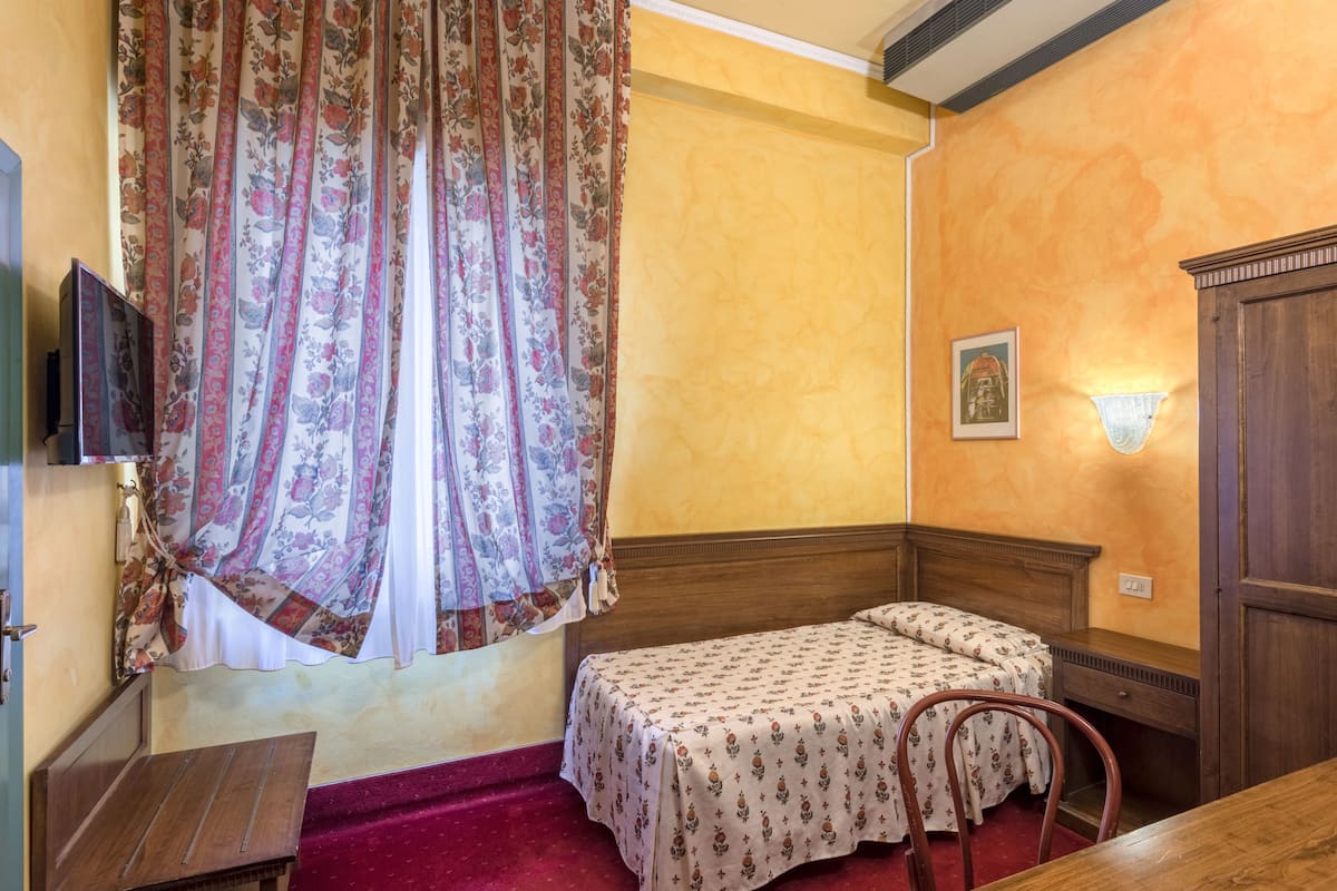 A small, cozy room with a single bed, a wooden nightstand, a small table, and a red carpet.