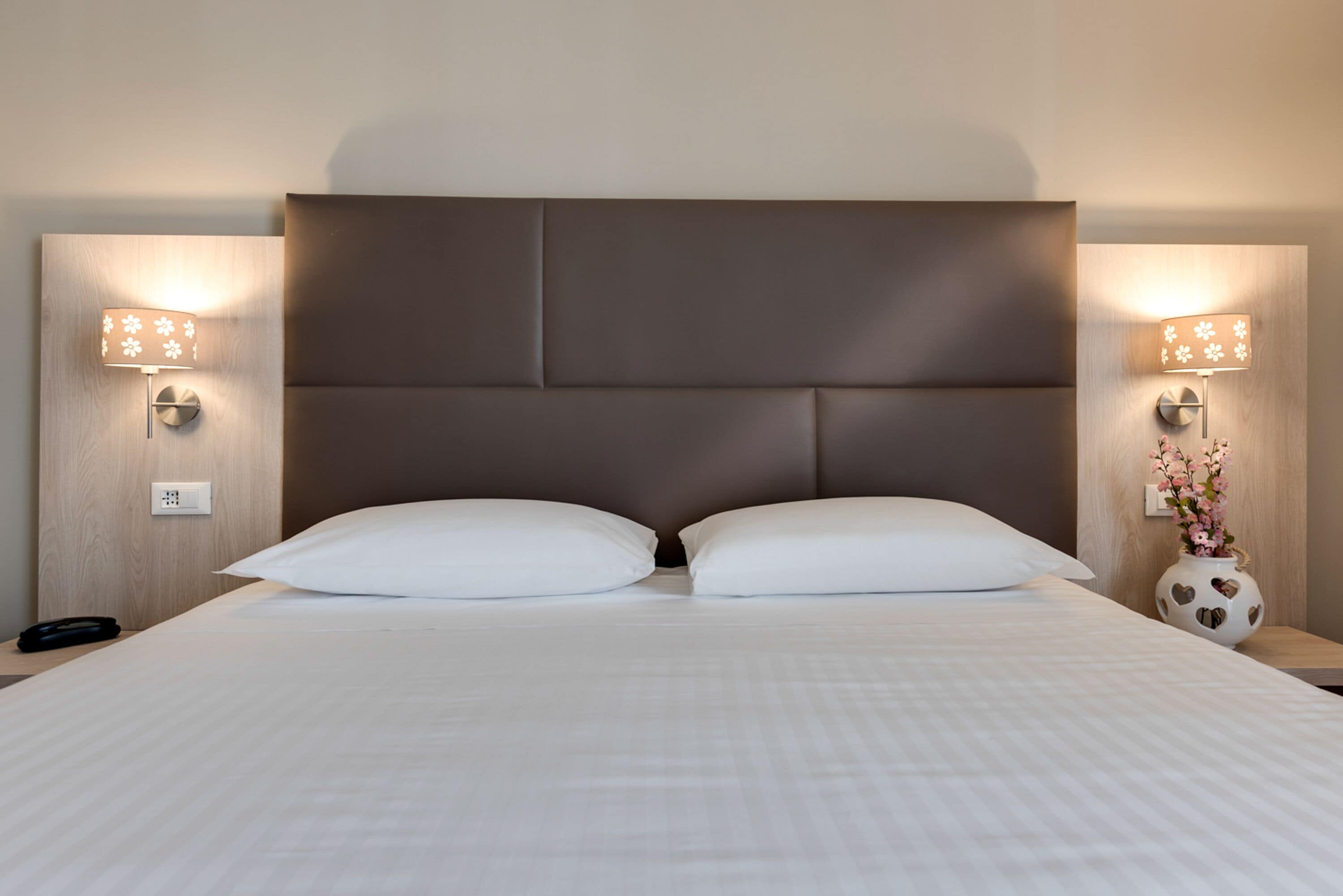 Uma cama bem arrumada com lençóis brancos, duas luminárias de cabeceira e um vaso decorativo com flores na mesa de apoio.