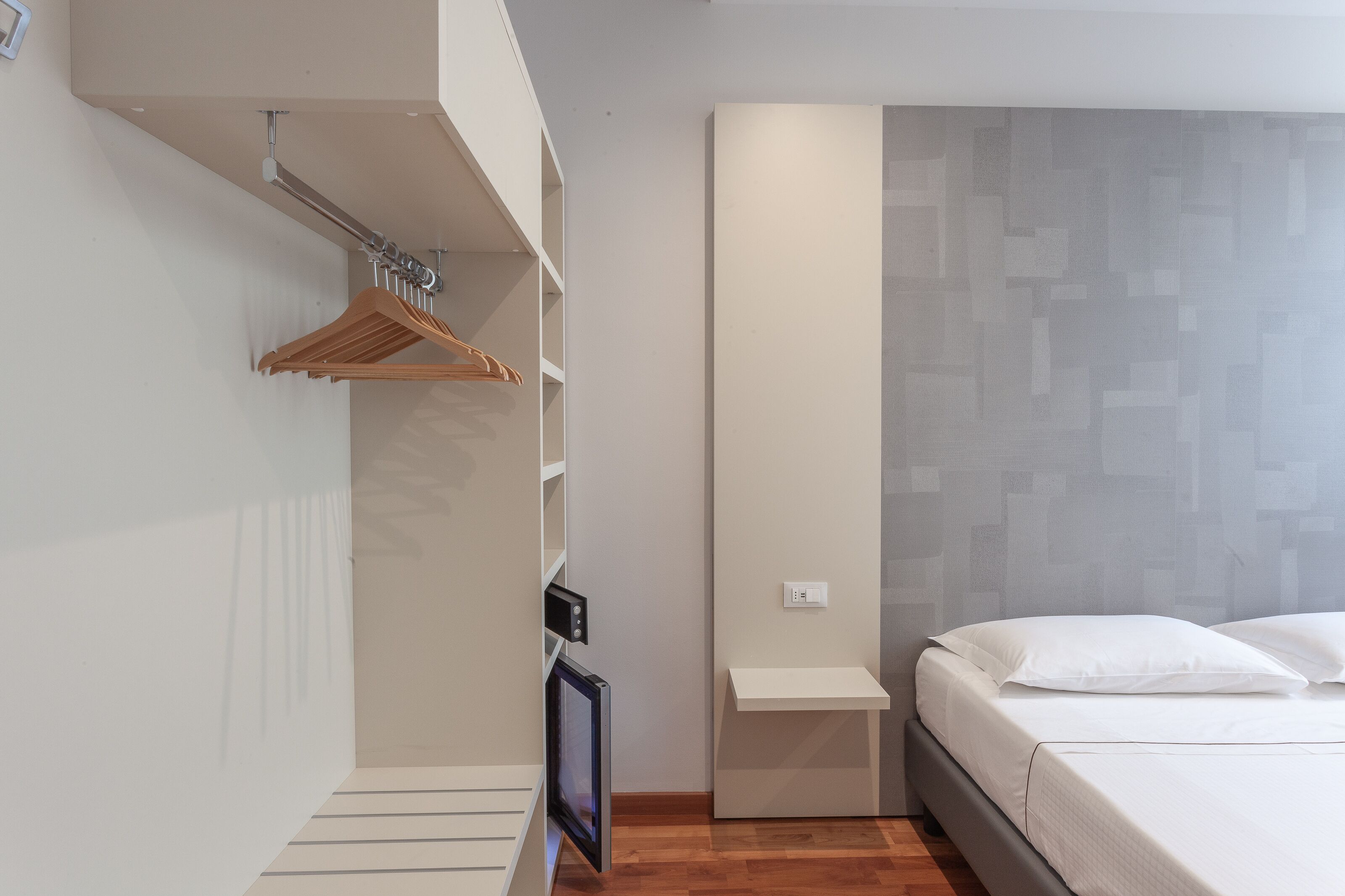 A bedroom with a bed, a bedside table, a hanging wardrobe, and a wall-mounted shelf.