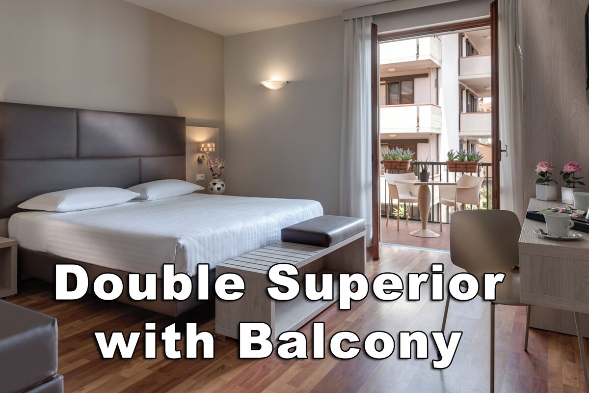 Habitación doble superior con balcón, que incluye una cama, mesitas de noche, un escritorio y vistas al área del balcón exterior.