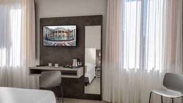 Une chambre d’hôtel comprenant un lit, un bureau avec une chaise, un téléviseur fixé au mur et une grande fenêtre avec des rideaux.