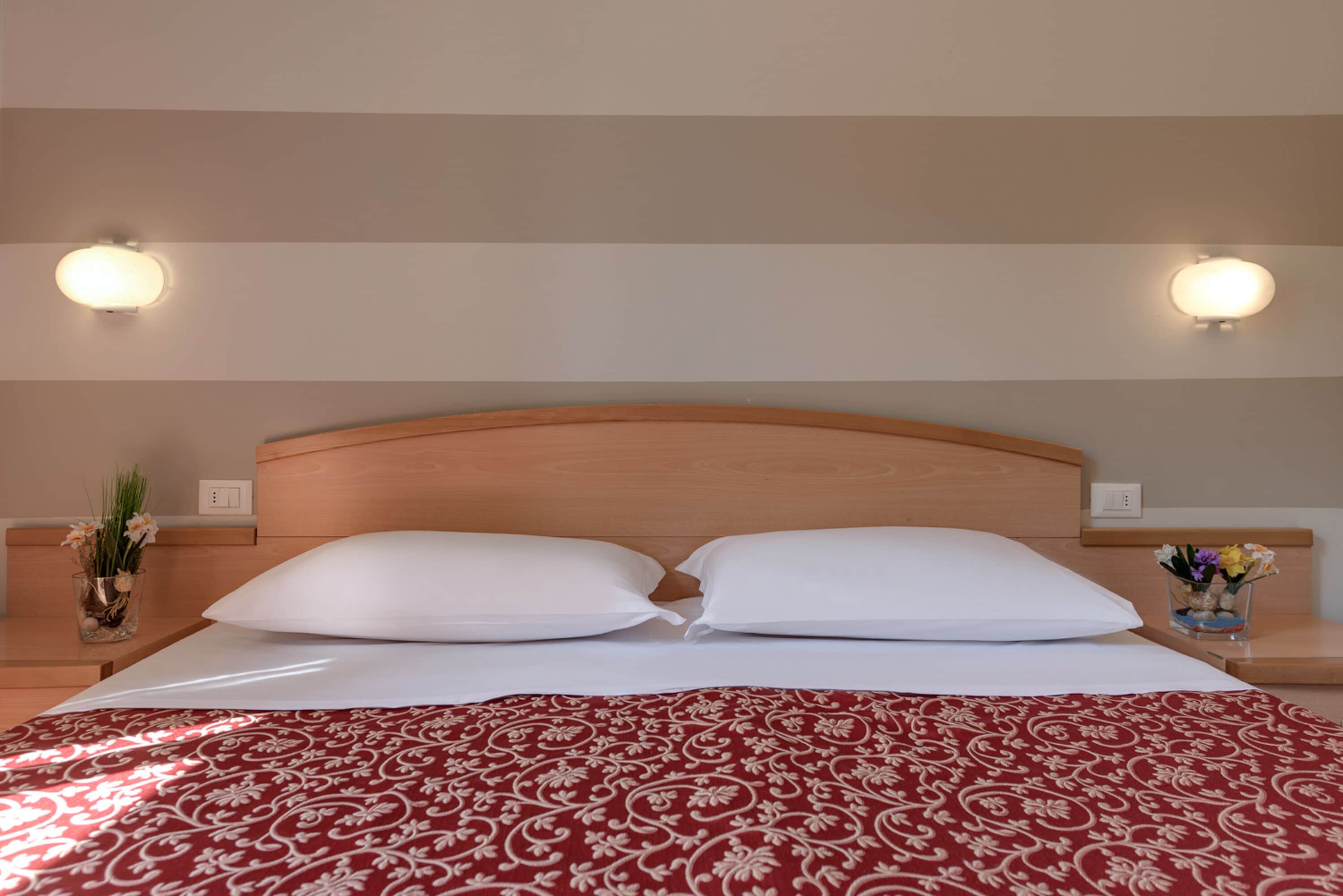 Habitación de hotel con cabecera de madera, dos almohadas blancas, colcha estampada roja y dos lámparas de pared.