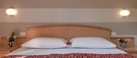 Habitación de hotel con cabecera de madera, dos almohadas blancas, colcha estampada roja y dos lámparas de pared.