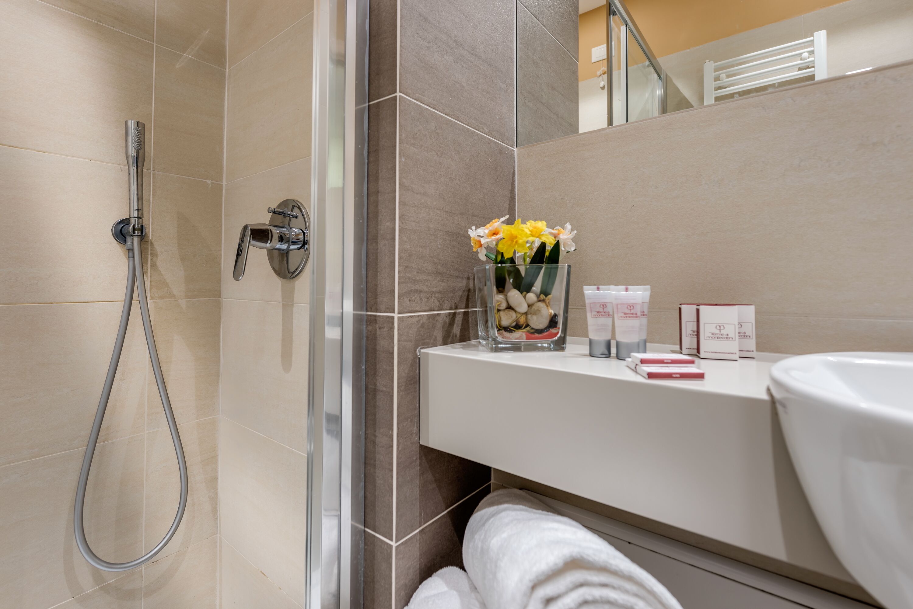 Un baño moderno con ducha de vidrio, tocador blanco con artículos de aseo y un jarrón con flores.
