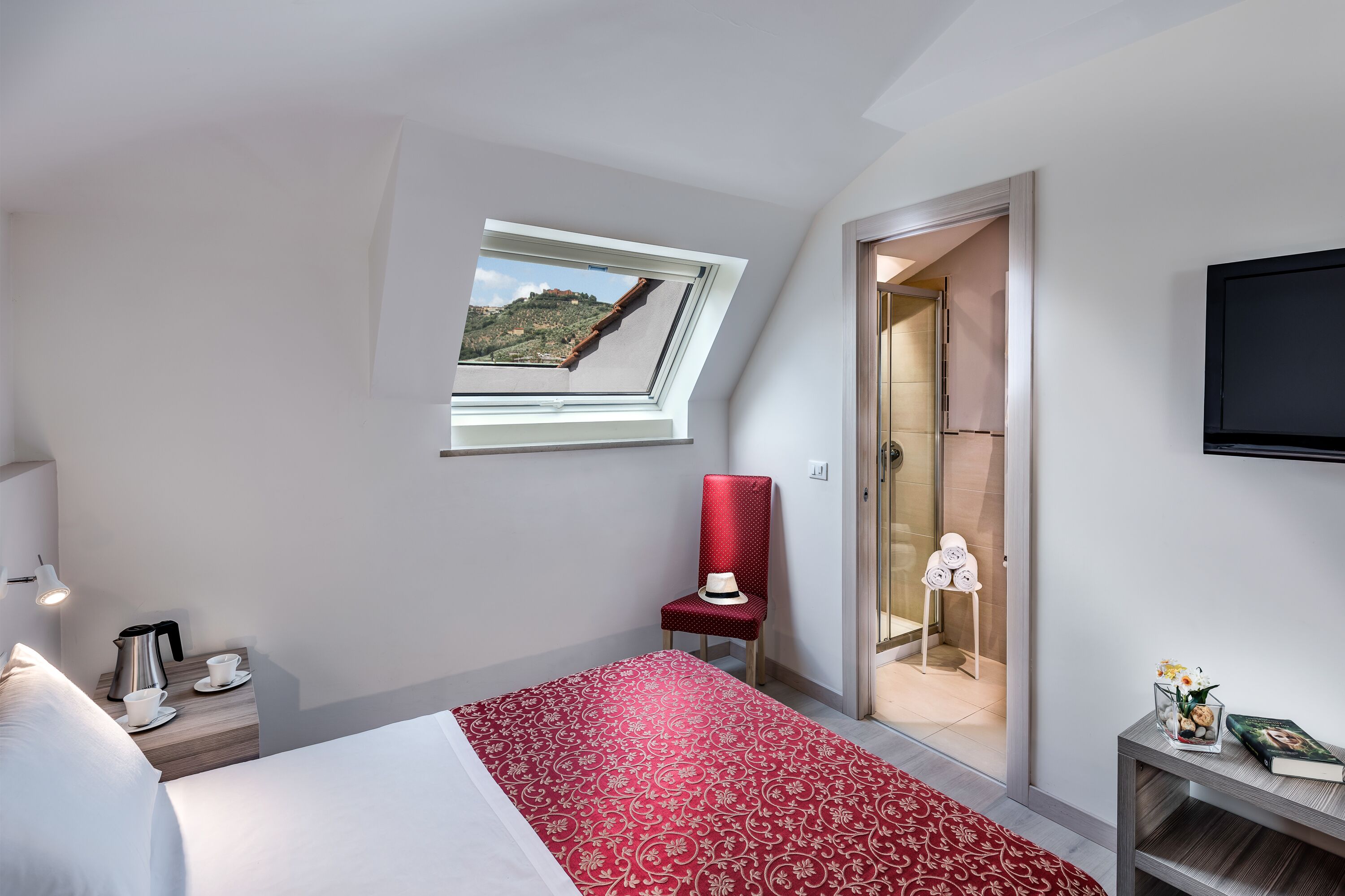 A hotel room with a bed, a red patterned bedspread, a red chair, a small table with a book and flowers, a television, and a window with a view of a hill.