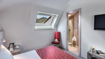 Hotelkamer met een bed, een rood beddegoed met patroon, een rode stoel, een klein tafeltje met een boek en bloemen, een televisie en een raam met uitzicht op een heuvel.