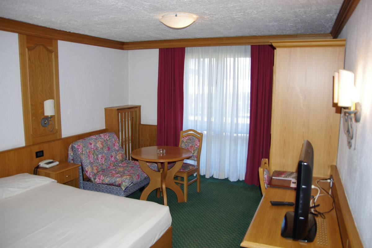 A hotel room with a bed, a desk with a computer, a chair, a small table, and a window with curtains.