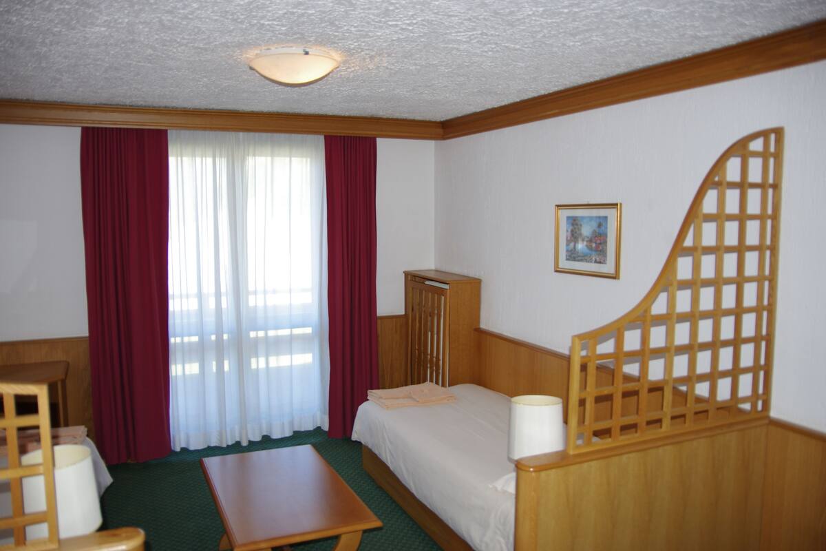 A hotel room with a bed, a wooden desk, a chair, a small table, a window with curtains, a picture on the wall, and a wooden partition.