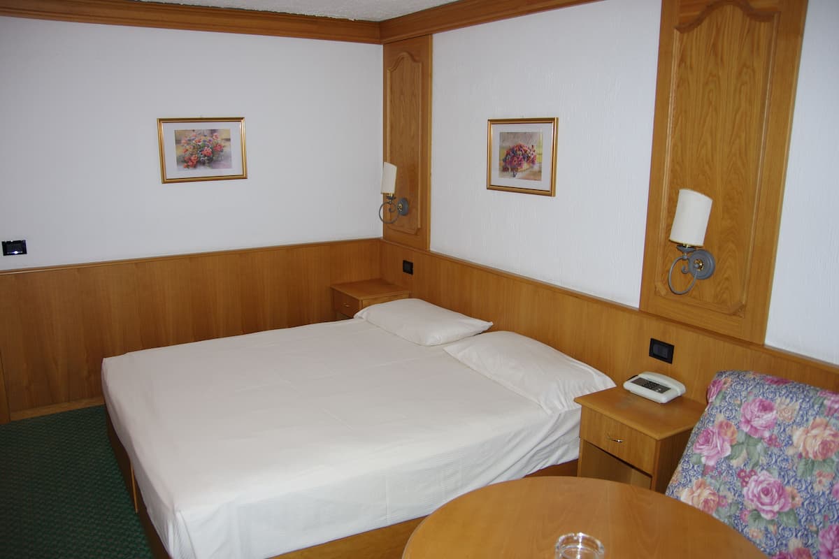 A hotel room with a single bed, a wooden headboard, two bedside tables with lamps, a small round table, and a floral chair.