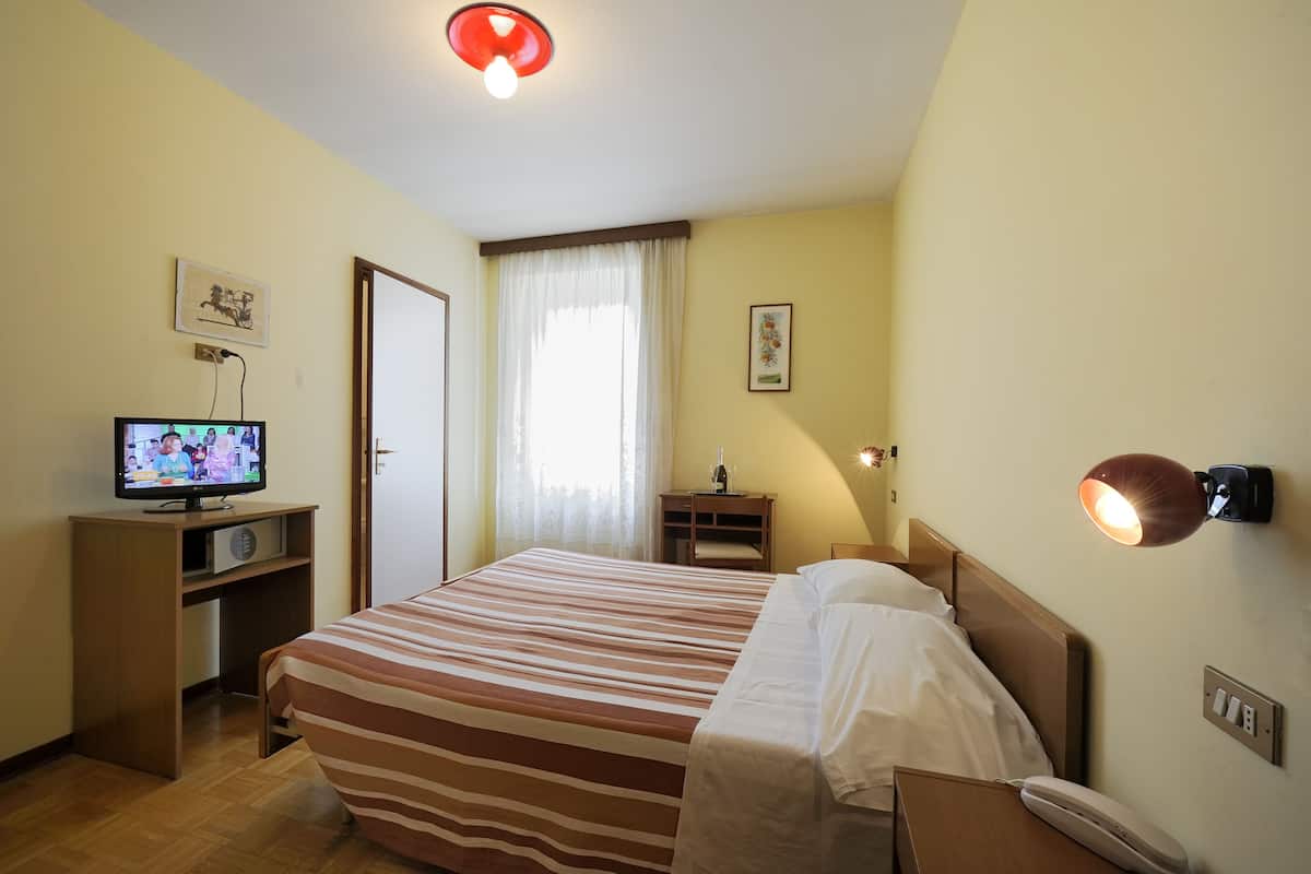 A hotel room with a bed, a TV on a stand, a bedside lamp, and a window with curtains.