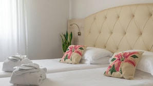 Habitación de hotel con dos camas, ropa de cama blanca, almohadas estampadas con motivos florales, una planta en la mesita de noche y una lámpara.