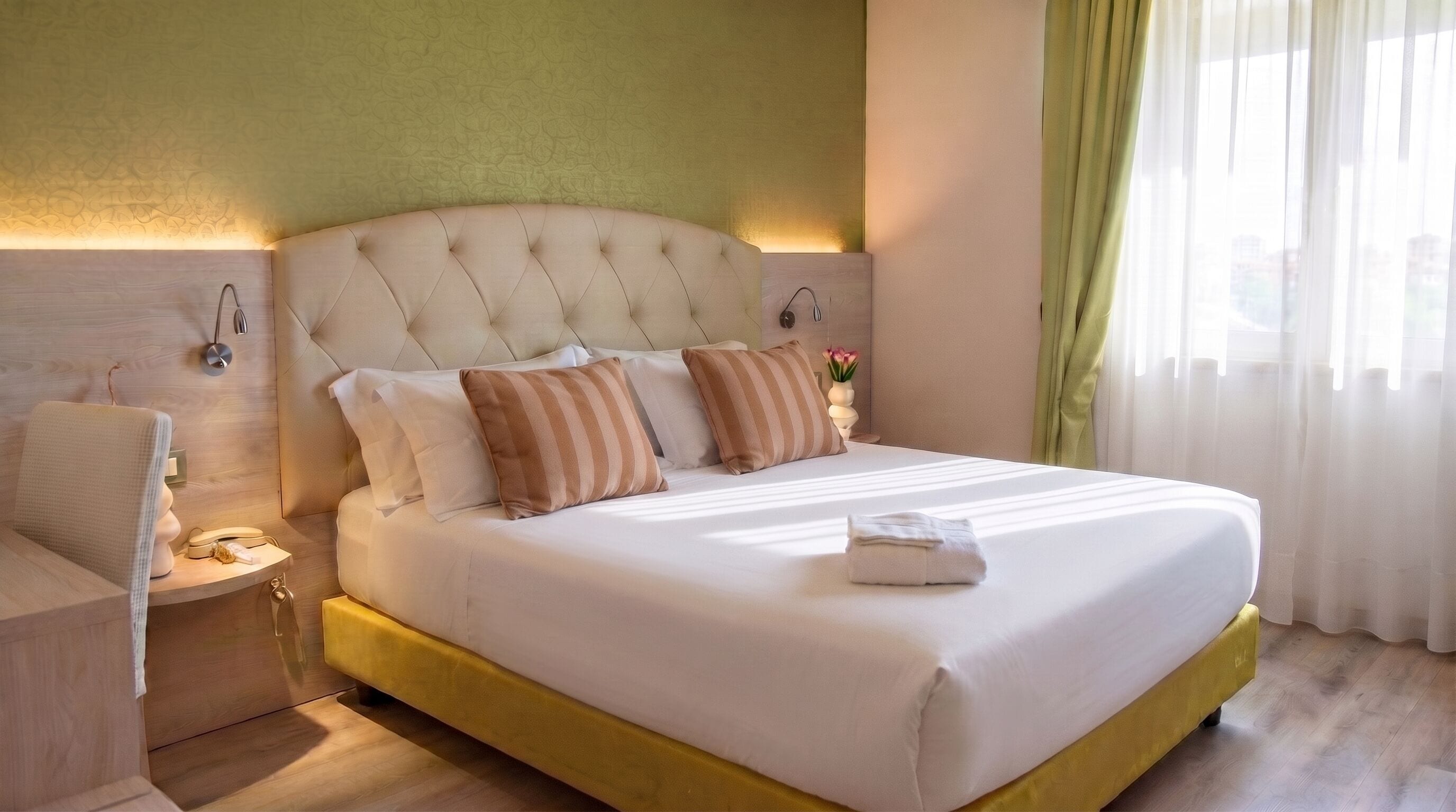 A neatly made bed with a tufted headboard, a bedside table with a lamp, and a window with sheer curtains.