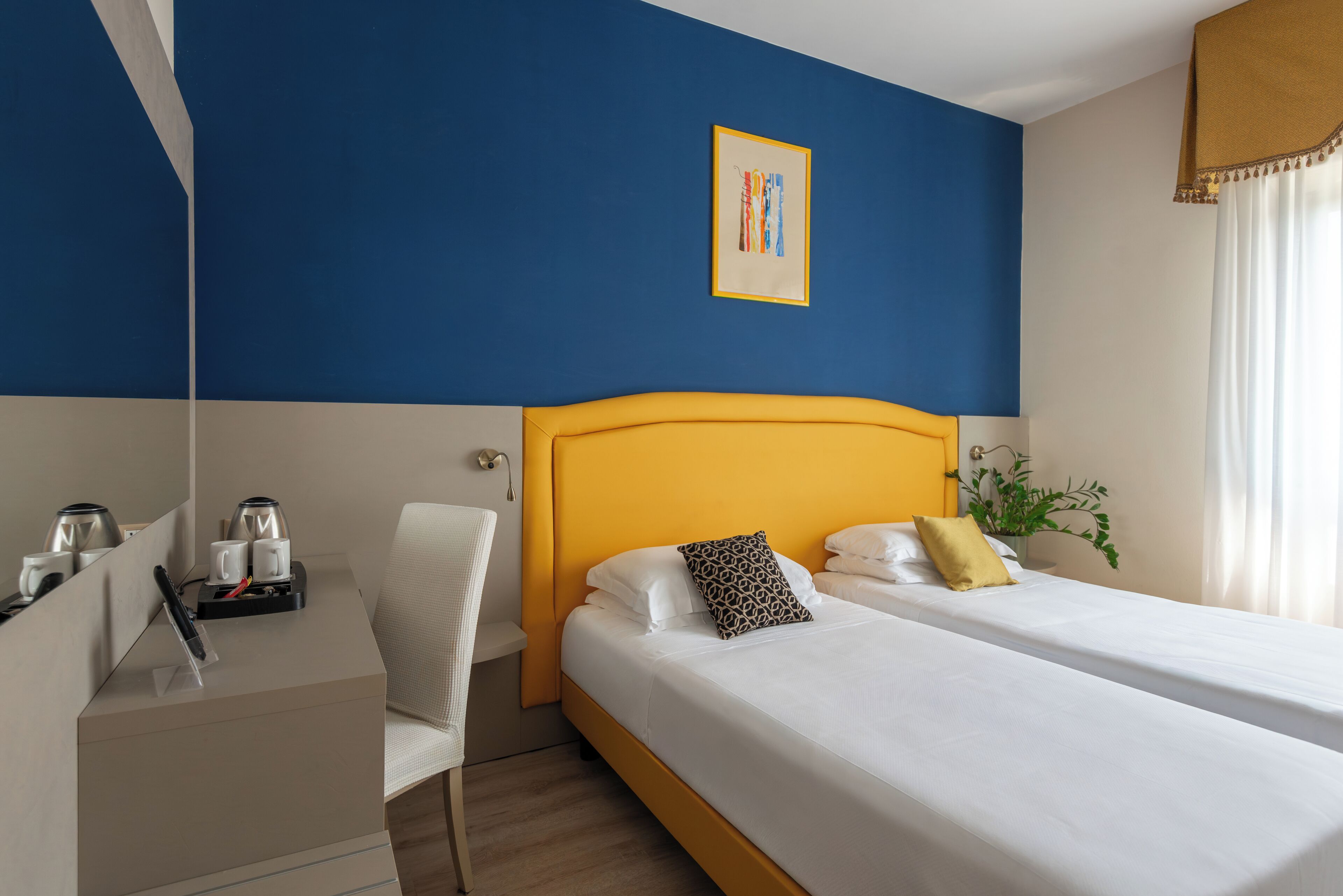 Ein Hotelzimmer mit gelbem Kopfteil, zwei Betten, einem Schreibtisch mit Stuhl und einer Kaffeemaschine.
