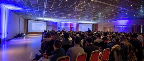 A conference room with rows of chairs facing a presentation screen, a stage, and a large audience.