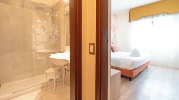 A hotel room with a glass-enclosed shower, a white sink, a small stool, a bed with a white mattress and orange headboard, and a window with sheer curtains.