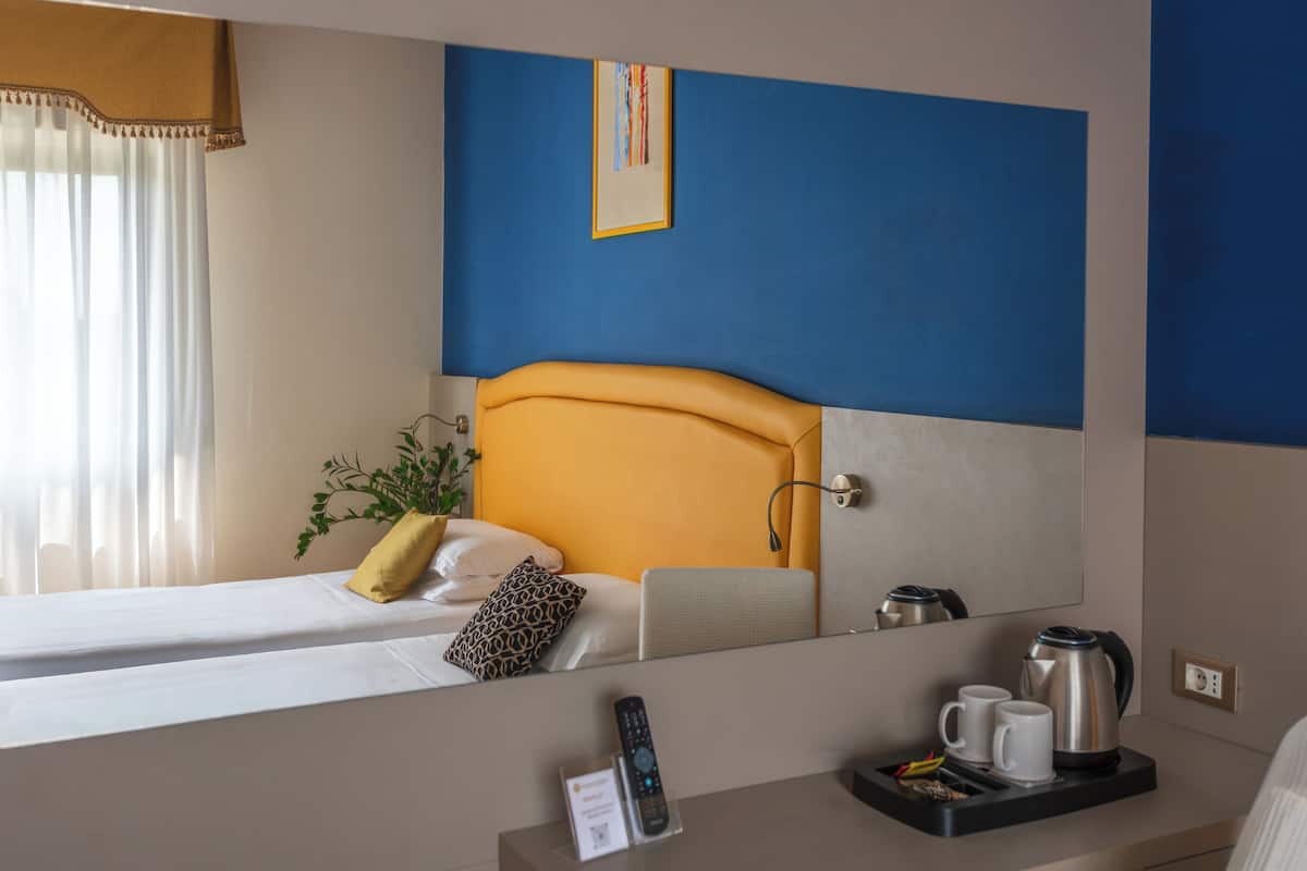 Ein Hotelzimmer mit einem Bett, einem Nachttisch mit Fernbedienung, einer Ablage mit Wasserkocher und Tassen sowie einem großen Spiegel, der das Zimmer reflektiert.