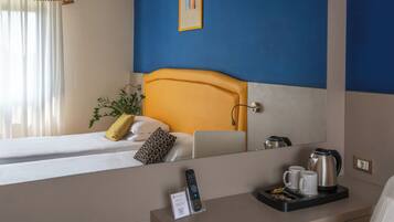 Una habitación de hotel con una cama, una mesita de noche con un control remoto, una bandeja con una tetera y tazas, y un espejo grande que refleja la habitación.
