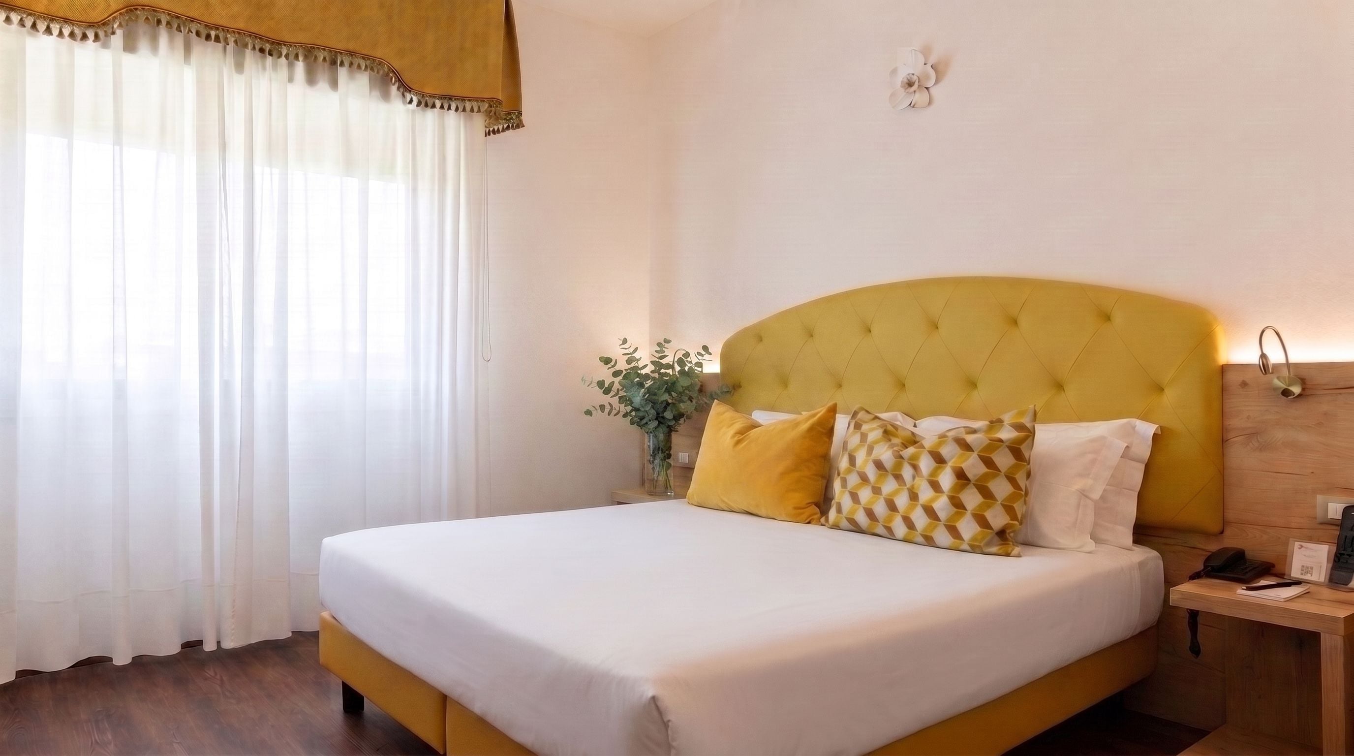 A hotel room with a bed, a nightstand, a vase with flowers, and a window with curtains.