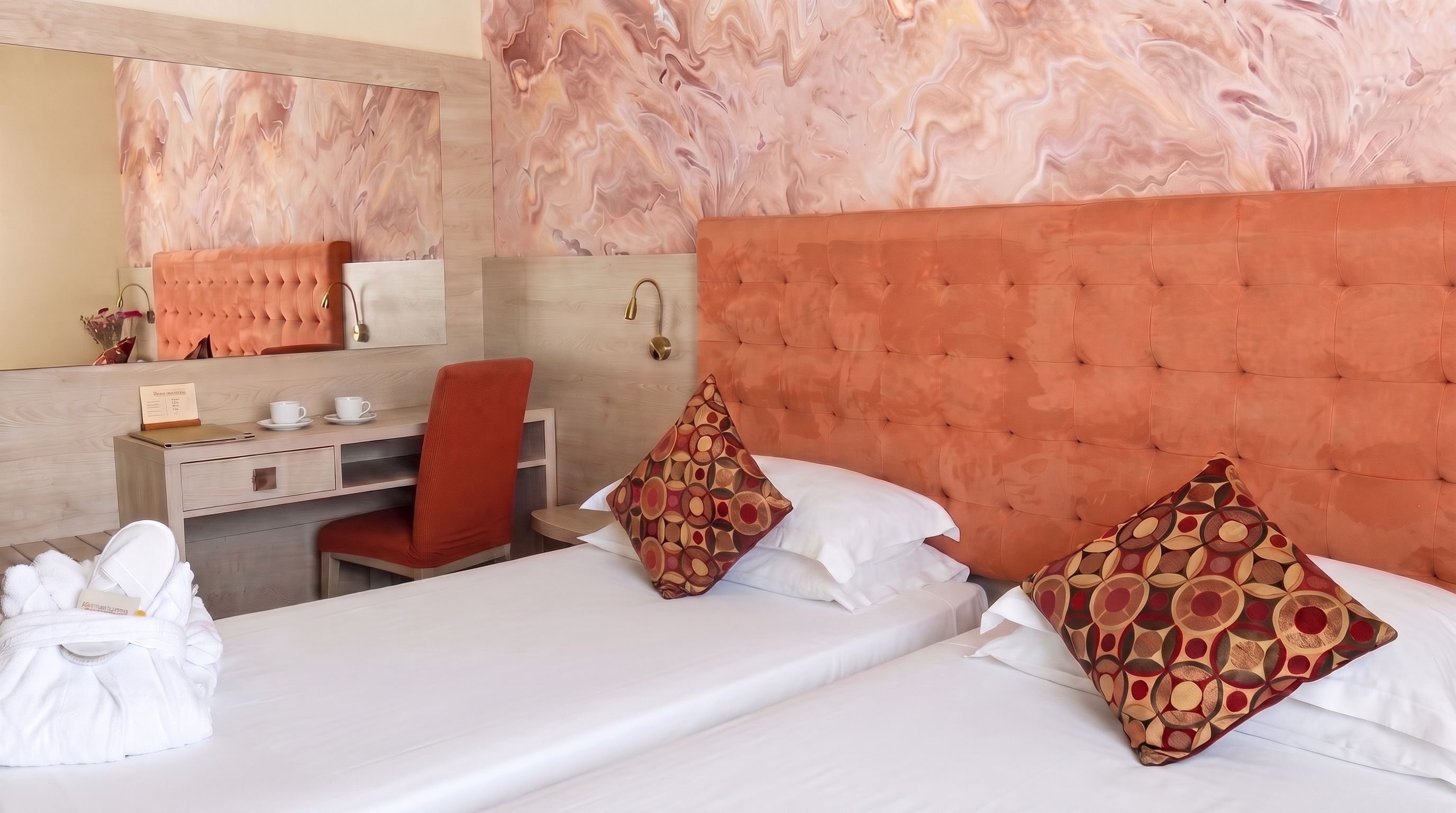 Habitació d'hotel amb un llit, dos almoinos, una taula de nit, una cadira, un escriptori, un mirall i una paret amb un dibuix semblant al marbre.